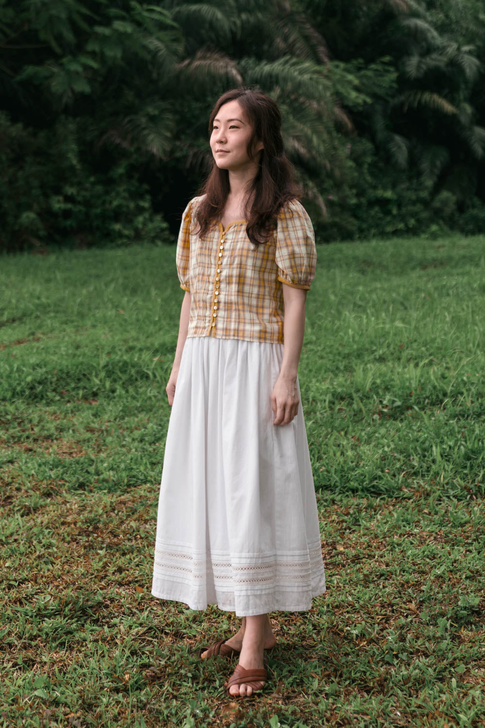 Woman in a plaid shirt and white skirt standing in a grassy field with trees in the background